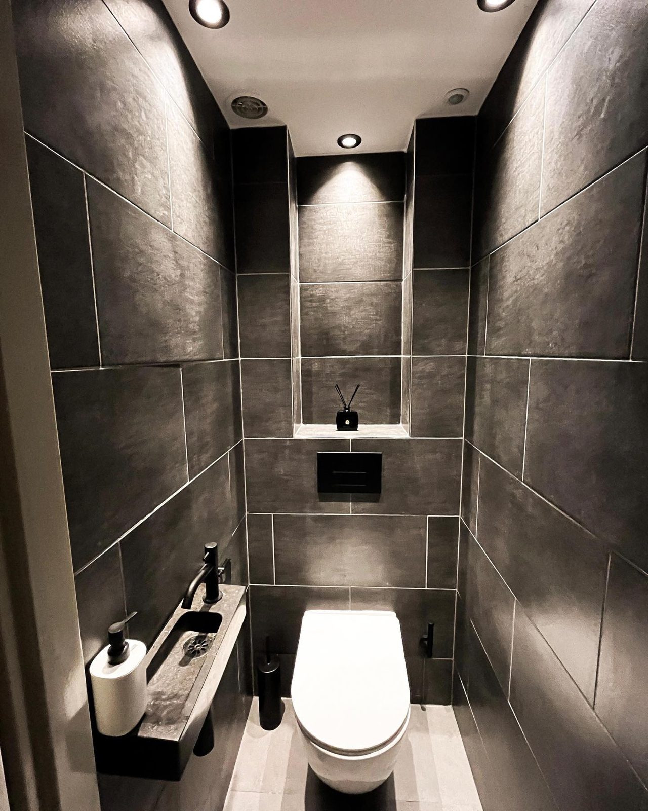 Gerenoveerd toilet met donkere tegels, hangtoilet en verlaagd plafond met spotjes